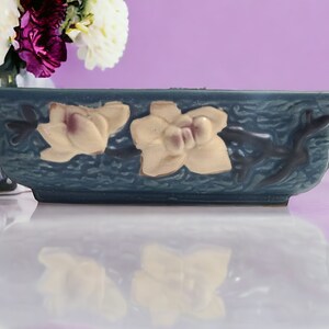 May include: A blue ceramic planter with a rectangular shape and two handles. The planter features a raised design of white flowers with purple centers.