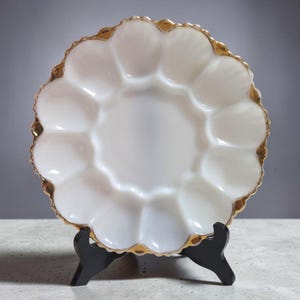 May include: A white, scalloped egg serving plate with a gold rim, displayed on a black stand. The plate has 12 individual egg compartments and a central serving area. The gold trim adds a touch of elegance to the vintage design.