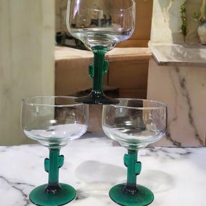 May include: Three clear glass margarita glasses with green cactus-shaped stems. The glasses are on a white marble surface.