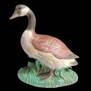 May include: A ceramic figurine of a brown and white goose standing on a green base. The goose has a long neck and a small head with a black eye.
