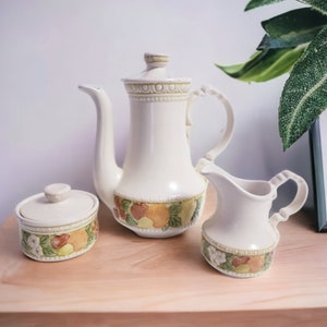 May include: A vintage ceramic tea set with a cream base and a floral pattern featuring fruits and leaves. The set includes a teapot, creamer, and sugar bowl.