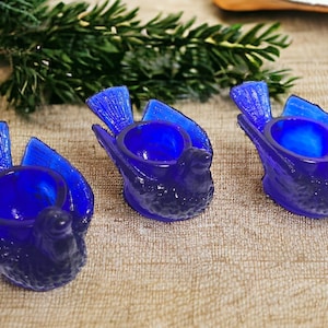 May include: Three blue glass bird-shaped egg cups. The cups are made of cobalt blue glass and have a detailed bird design.