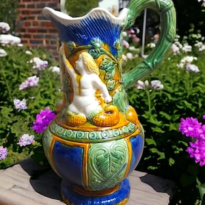 May include: A blue and yellow ceramic pitcher with a green handle and a white mermaid figure. The pitcher has a detailed design with leaves and geometric patterns.