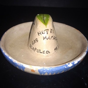 May include: A small, white ceramic sombrero with blue trim and a green leaf on top. It says "Hotel Las Hamacas Acapulco, Mex." on the side.