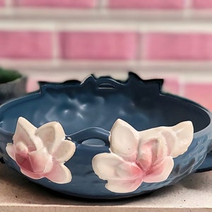 May include: A blue ceramic bowl with two white and pink magnolia flowers on the sides. The bowl has a scalloped edge and two handles.