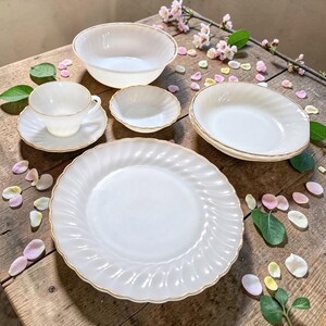 May include: A collection of white dishware with gold trim, including a large bowl, a small bowl, a teacup and saucer, and a dinner plate. The pieces are arranged on a wooden surface with floral accents.