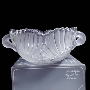 May include: A clear glass serving bowl with two handles. The bowl has a scalloped edge and a decorative pattern of overlapping fan shapes. The bowl is sitting on a blue box with the text "Kristallglas Crystal Glass Cristallin" printed on it.