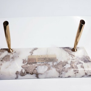 May include: A white marble pen holder with two gold pen slots. A small silver plaque with the text "Jim Newman" is attached to the front of the holder.