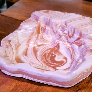 May include: A white and brown ceramic ashtray with a swirled pattern. The ashtray is shaped like a rock and has a deep well for holding ashes.