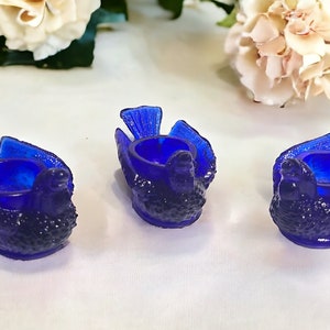 May include: Three cobalt blue glass bird-shaped egg cups. Each cup is shaped like a bird with its wings spread out. The cups are made of a textured glass and have a glossy finish.