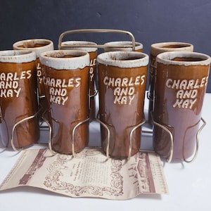 May include: Set of six brown ceramic mugs with white lettering that reads "Charles and Kay". The mugs are in a gold metal stand.