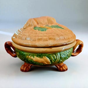 May include: A ceramic lidded serving dish with a tan, textured top and green base. The lid features a sculpted dog, and the base has a relief pattern. The dish has two handles and rests on three paw-shaped feet. A decorative kitchenware item.