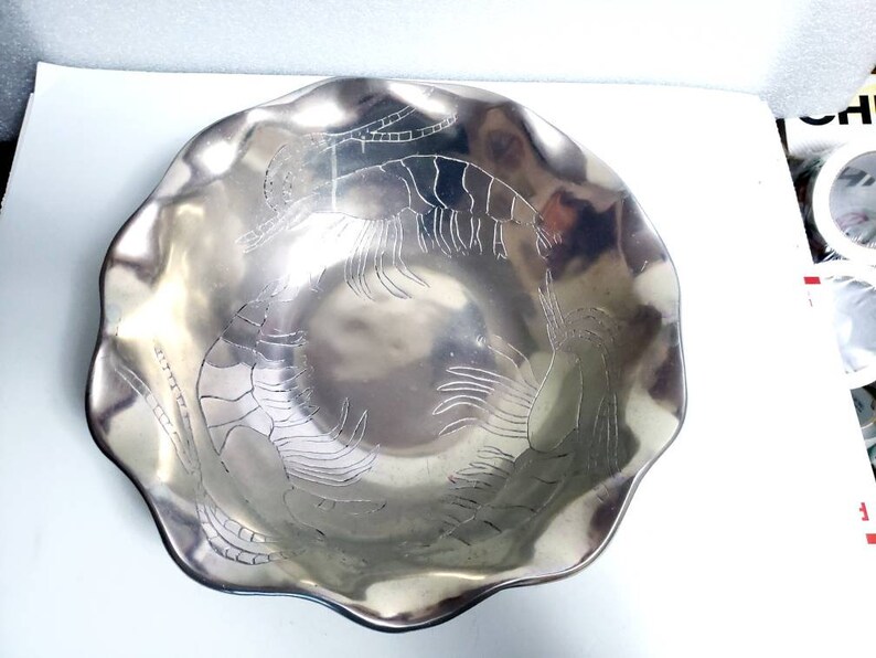 Peut inclure: Un bol de service en argent avec un bord festonn&eacute; et un motif en relief de deux crevettes. Le bol est en m&eacute;tal et a une finition brillante.