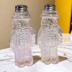 May include: Two vintage salt and pepper shakers in the shape of bearded men. The clear glass shakers have silver metal lids and are tinted with a soft pink hue. The figures are wearing hats and have detailed facial features.