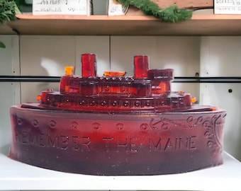 以下が含まれることがあります： 「Remember the Maine」という文字が刻まれた赤いガラス製の船の彫刻。彫刻は白い棚の上に置かれている。