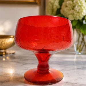 May include: A vibrant red glass bowl featuring a distinct crackle texture throughout the surface. The bowl is supported by a sturdy pedestal base and a circular foot, creating a stemmed silhouette that elevates the wide, rounded basin.