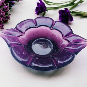 May include: A purple glass bowl shaped like a flower with a ruffled edge. The bowl is made of clear glass with a purple tint and has a small, round center.