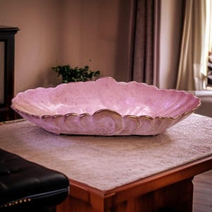 May include: A large, oval-shaped pink ceramic serving bowl with a scalloped rim and gold trim. The bowl has a speckled texture and a decorative swirl design at each end. A vintage-style decorative piece.