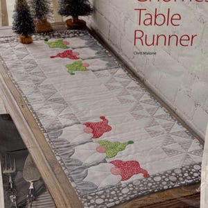 May include: A quilted table runner featuring a repeating pattern of gnomes with red and green polka-dot hats. The runner has a white center, gray geometric borders, and a gray polka-dot edge. Text on the image reads Gnomes Table Runner Chris Malone.