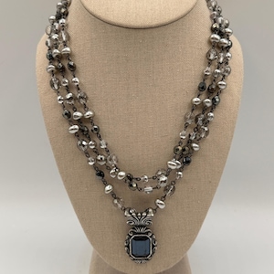 May include: A three-strand necklace with gray and clear beads and a square pendant. The pendant has a dark blue stone and an ornate silver-tone frame. The necklace is displayed on a beige mannequin.