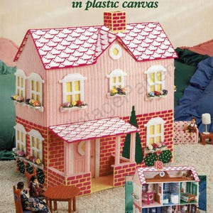 May include: A craft book titled My Pink Dollhouse in plastic canvas by Karen McDanel, featuring a large pink dollhouse with a white roof, brick-patterned base, and window boxes with flowers, plus a smaller inset image showing the interior rooms.