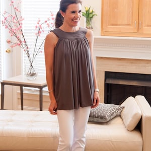 May include: A woman wearing a gray sleeveless top with a gathered neckline and white pants. She is standing in front of a white couch with a gray pillow.