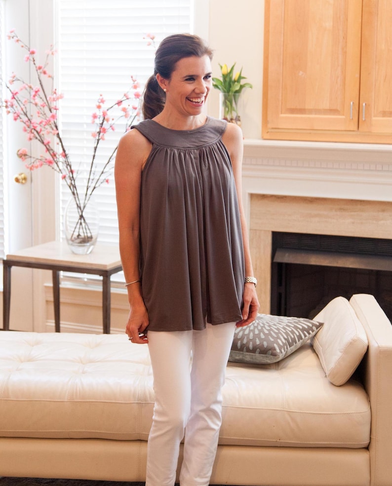 May include: A woman wearing a gray sleeveless top with a gathered neckline and white pants. She is standing in front of a white couch with a gray pillow.