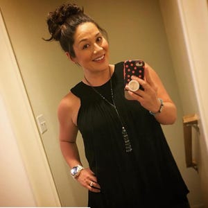 May include: A woman with brown hair in a bun is wearing a black sleeveless top with a long beaded necklace. She is holding a black and pink polka dot phone case with a white pop socket.