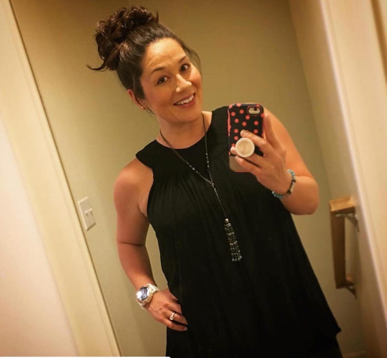 May include: A woman with brown hair in a bun is wearing a black sleeveless top with a long beaded necklace. She is holding a black and pink polka dot phone case with a white pop socket.