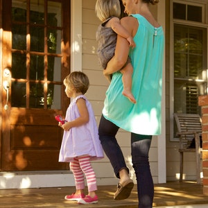 May include: A woman in a turquoise sleeveless top carries a young child in her arms while walking on a porch. A young girl in a lavender dress and pink and white striped leggings walks beside them.