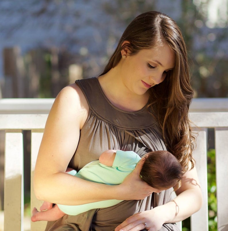 May include: A woman in a brown nursing top is holding a baby in her arms. The baby is wearing a light blue onesie and is sleeping. The woman is looking down at the baby.