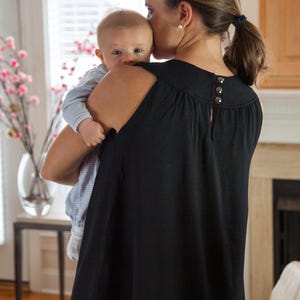 May include: A woman wearing a black sleeveless top with a scoop neck and a button detail on the back is holding a baby in her arms. The baby is wearing a blue and white striped shirt. The woman is looking down at the baby.