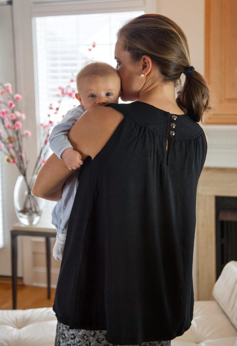 May include: A woman wearing a black sleeveless top with a scoop neck and a button detail on the back is holding a baby in her arms. The baby is wearing a blue and white striped shirt. The woman is looking down at the baby.