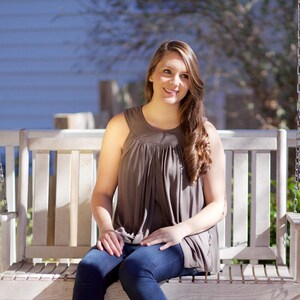 May include: A woman sits on a white wooden swing, wearing a brown sleeveless top and blue jeans. She is smiling and looking to the side.
