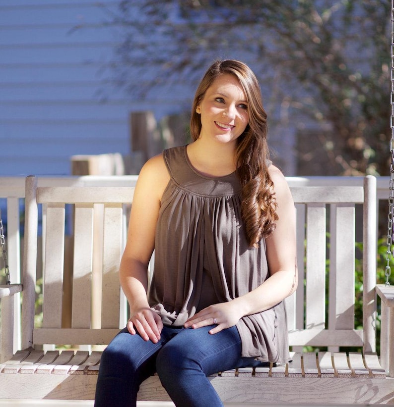 May include: A woman sits on a white wooden swing, wearing a brown sleeveless top and blue jeans. She is smiling and looking to the side.