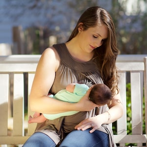 May include: A woman wearing a brown sleeveless top sits on a white porch swing and nurses a baby wearing a light blue shirt.