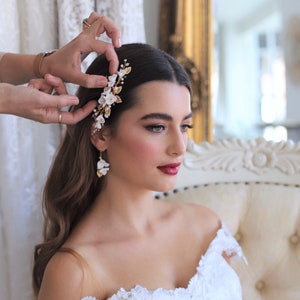 May include: A bride with long brown hair wearing a white floral hair comb and matching earrings. The comb has gold accents and white flowers. The bride is wearing a white off-the-shoulder dress.