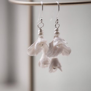 May include: White flower earrings with pearl accents. The earrings are made of delicate, layered petals and hang from silver hooks.