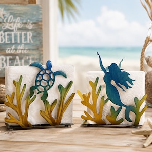 May include: Two metal napkin holders with a beach theme. One features a blue sea turtle, the other a blue mermaid. Both have yellow and green coral accents. The holders sit on a wooden surface with a beach scene in the background.