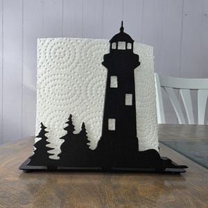 May include: Black metal lighthouse-shaped paper towel holder. The holder features a silhouette of a lighthouse with windows and a detailed top, along with a forest scene at the base. The holder is designed to hold a roll of paper towels.