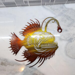 May include: A metal wall art sculpture of a anglerfish with a long, thin, curved lure. The fish is a reddish-brown color with a yellow-gold head and has a large, toothy mouth. The sculpture is on a white marble background.