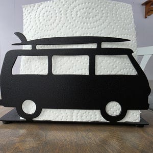 May include: Black metal van-shaped paper towel holder with a surfboard on top. The holder has a rectangular base and holds a roll of white paper towels. The design is a silhouette of a classic van.