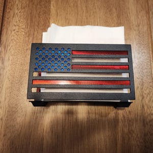 May include: A black metal napkin holder shaped like the American flag. The holder has red and blue stripes and a blue field of stars. The holder is mounted on a wooden surface.