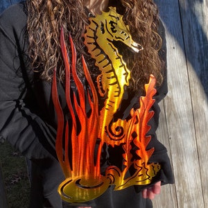 May include: A metal seahorse sculpture with a gradient of orange and yellow paint. The seahorse is standing in a bed of seaweed.