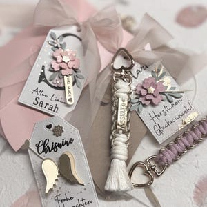 May include: Three rectangular gift tags with ribbons and two macrame keychains. One tag features a pink flower and 'Alles Liebe Sarah', another has a pink flower and 'Herzlichen Glückwunsch', and the third has gold wings and 'Frohe Weihnachten'.