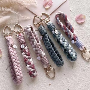 May include: Six braided cord keychains with gold-tone heart-shaped clasps. The keychains feature various color combinations including solid pink, multi-toned pink, grey, and blue. Several keychains include small circular charms with letters M, E, A, S, D, and J.