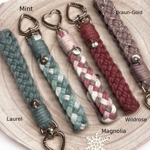 May include: Five braided cord keychains with heart-shaped metal clasps. Colors include Laurel, Mint, Magnolia, Wildrose, and Braun-Gold. Each keychain features a small circular metal charm with a letter: X, M, W, E, and a blank charm.