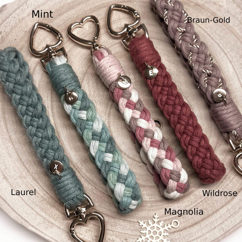 May include: Five braided cord keychains with heart-shaped metal clasps. Colors include Laurel, Mint, Magnolia, Wildrose, and Braun-Gold. Each keychain features a small circular metal charm with a letter: X, M, W, E, and a blank charm.