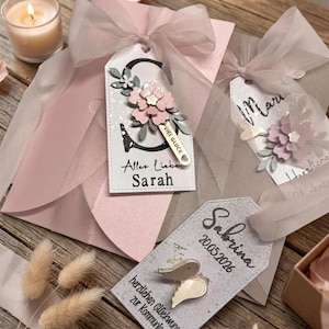 May include: Three rectangular gift tags with rounded tops, each featuring a sheer ribbon bow. One tag displays the letter S, a pink flower, and the text Alles Liebe Sarah. Another shows the name Maria and a flower. The third shows Sabrina, 20.05.2026, and wings.