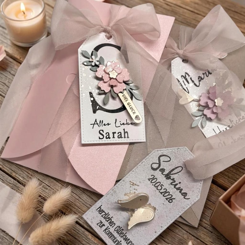 May include: Three rectangular gift tags with rounded tops, each featuring a sheer ribbon bow. One tag displays the letter S, a pink flower, and the text Alles Liebe Sarah. Another shows the name Maria and a flower. The third shows Sabrina, 20.05.2026, and wings.